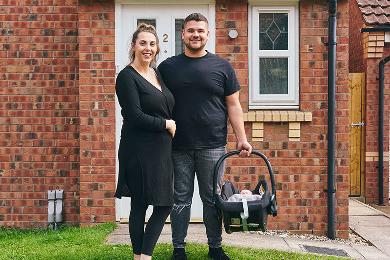 Couple standing in front of their new home with a baby in a carrier.