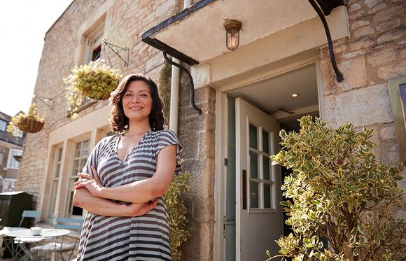 Woman smiling outside a charming stone house with greenery and flowers.
