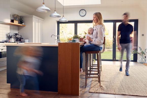 Mother sitting at a kitchen island with a baby, while a child runs by and a man walks in.